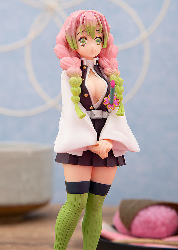 An anime collectible figurine from Demon Slayer, styled with pink hair, a white haori jacket, dark pleated skirt, and green socks, set against a soft-focus background.